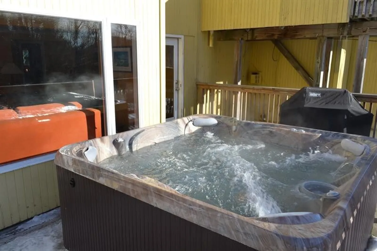 This exterior image showcases the hot tub in a different season, highlighting the surrounding outdoor seating area and a glimpse through windows into the retreat's interior. The setup suggests flexibility for year-round use and a social, tranquil environment.