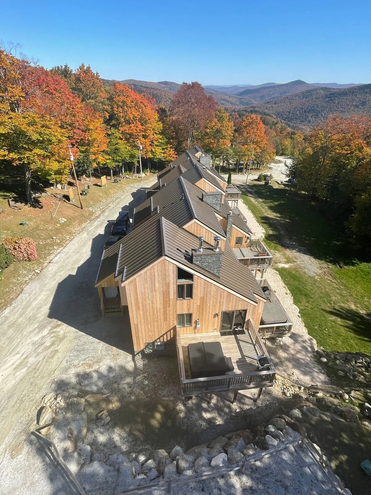 A wide view of vibrant autumn foliage surrounding rustic wooden cabins, with a dramatic mountain backdrop. The scene includes well-maintained outdoor spaces ideal for hiking, stargazing, and enjoying Vermont's natural beauty. A private deck is visible, offering a space to relax and soak in the scenery.