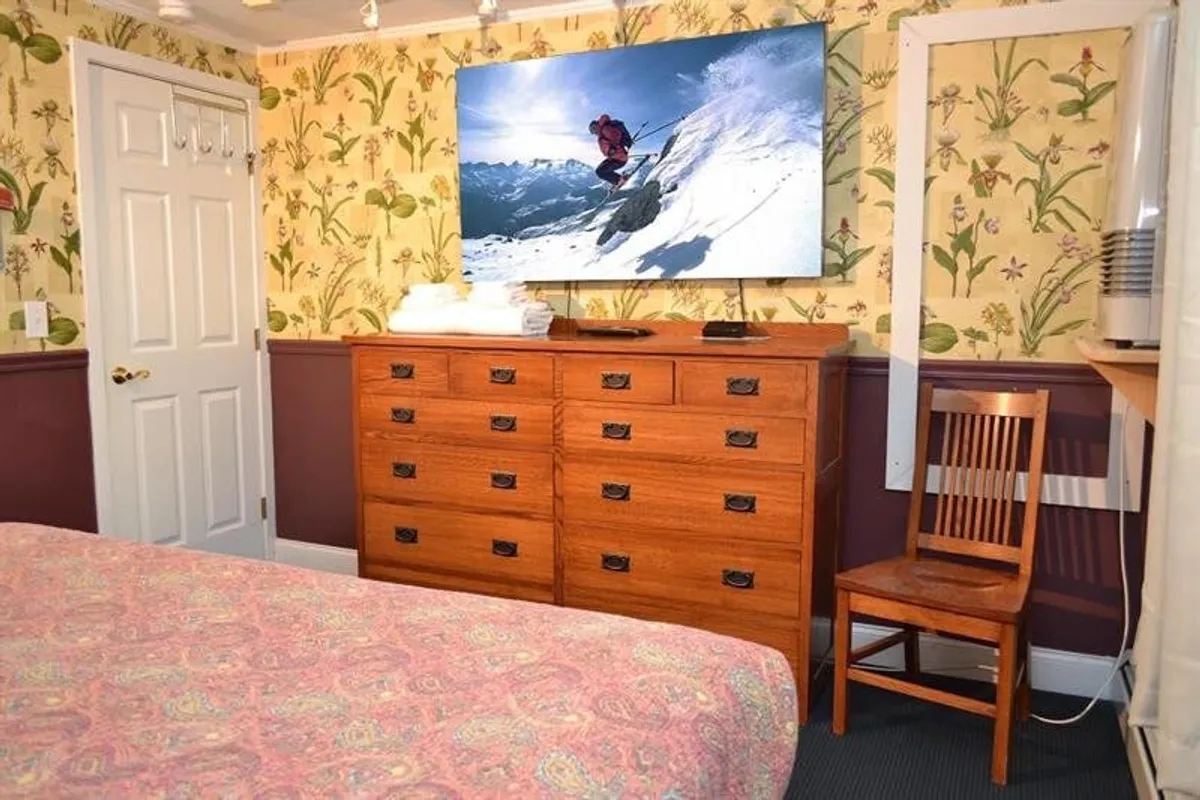 This image captures a comfortable interior with a large television displaying a skiing scene, a wooden dresser, and floral wallpaper. The overall scene suggests a personalized, homey environment suitable for relaxation and extended stays.
