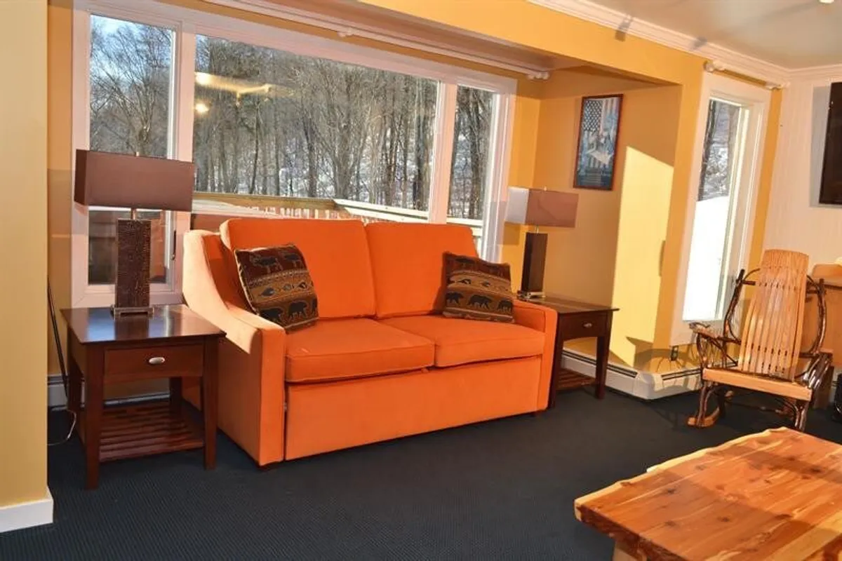 A cozy, inviting living area features an orange sofa, large windows letting in abundant natural light, a wooden coffee table, and a rocking chair. The scene emphasizes relaxation, with the opportunity to enjoy morning coffee or unwind while watching the sunset.