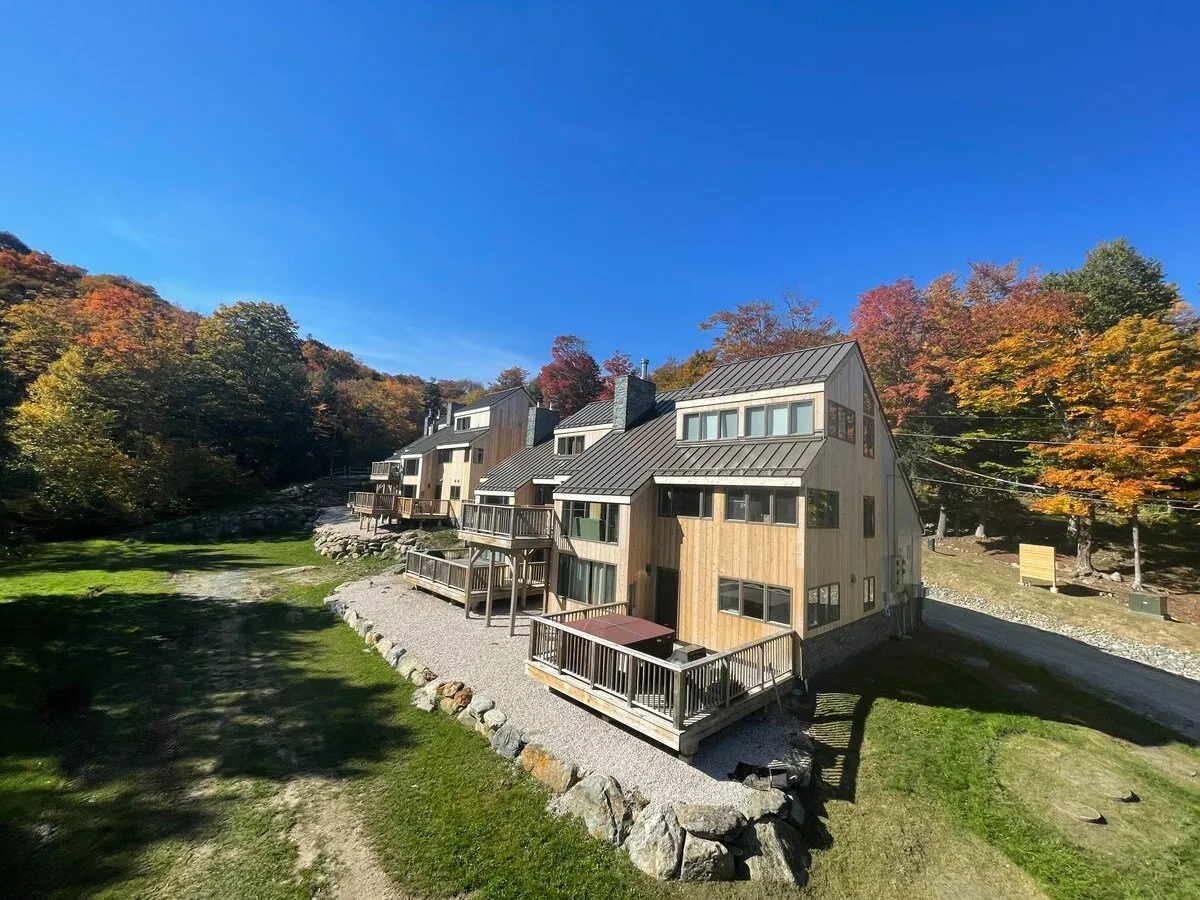 This exterior image features a cluster of modern wooden houses nestled among colorful autumn foliage beneath a clear blue sky. The scene suggests outdoor leisure, including relaxing on the deck and stargazing, with a sense of tranquility and connection to nature.