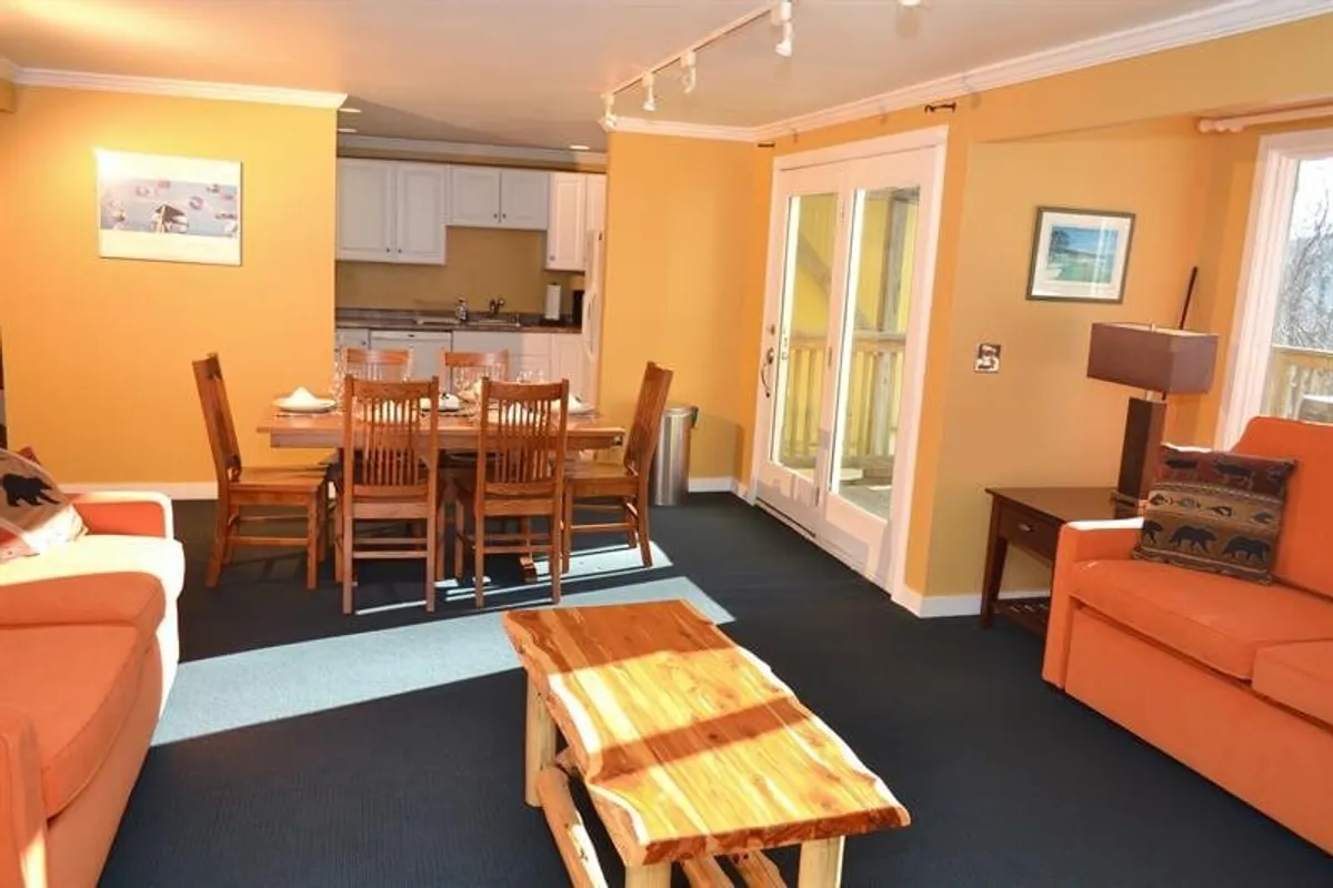 This image depicts a bright, welcoming living and dining area centered around a dining table, orange sofa, and wooden coffee table, with double doors that open to the outside. The scene suggests family gatherings, shared meals, and easy transitions between indoor comfort and outdoor adventure.