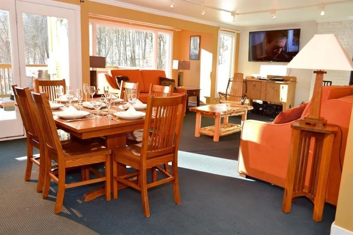 The image showcases a warm, inviting living and dining area with a cozy sofa, a dining table set for gatherings, a television for entertainment, and large windows that likely let in beautiful natural light. The lighting from the lamps creates an intimate, comfortable atmosphere, perfect for relaxing after a day on the slopes.