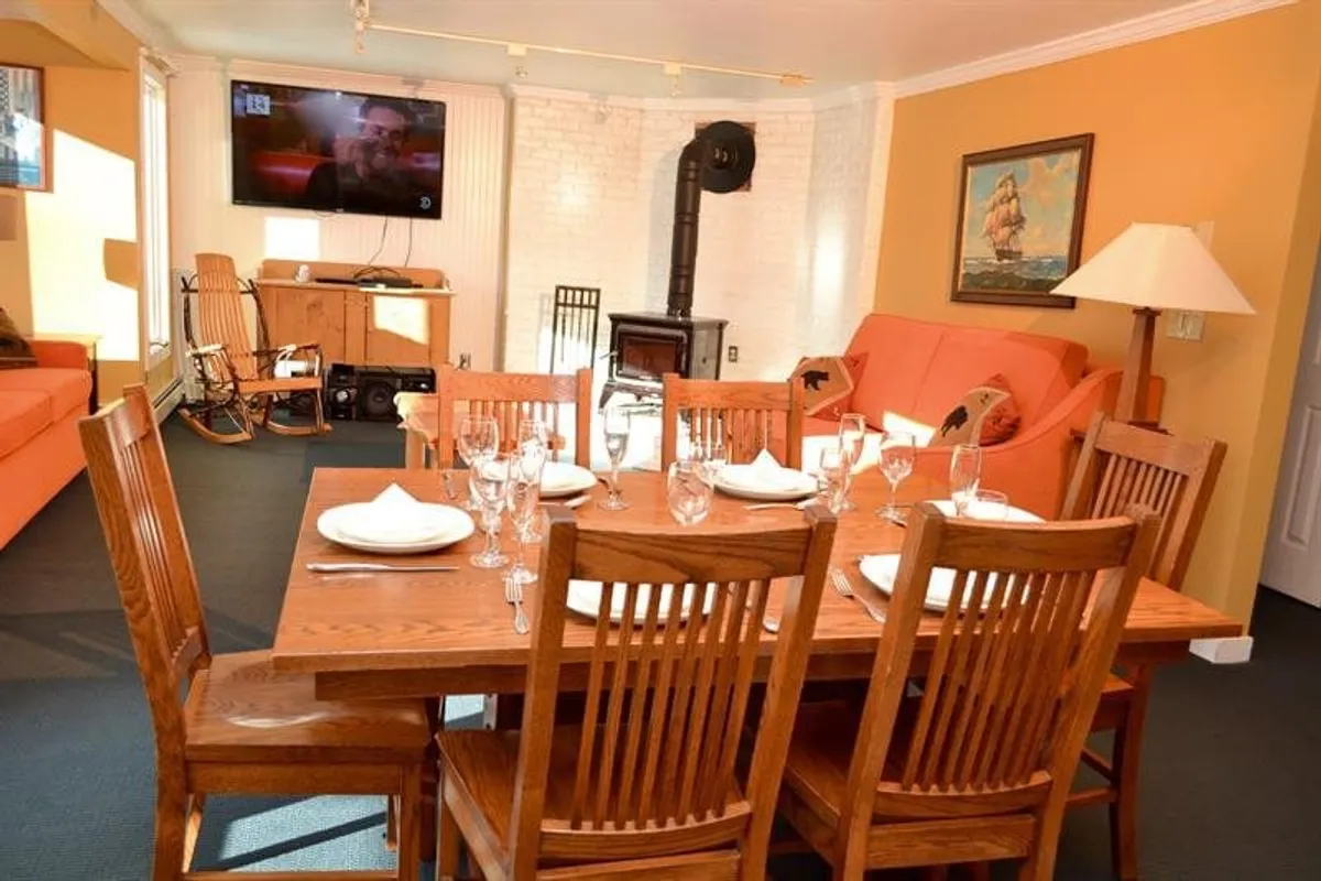This image features a cozy, open-plan living and dining area. It shows a wood stove as a focal point, a sofa for lounging, a dining table for shared meals or board games, and a television—all bathed in warm natural light.