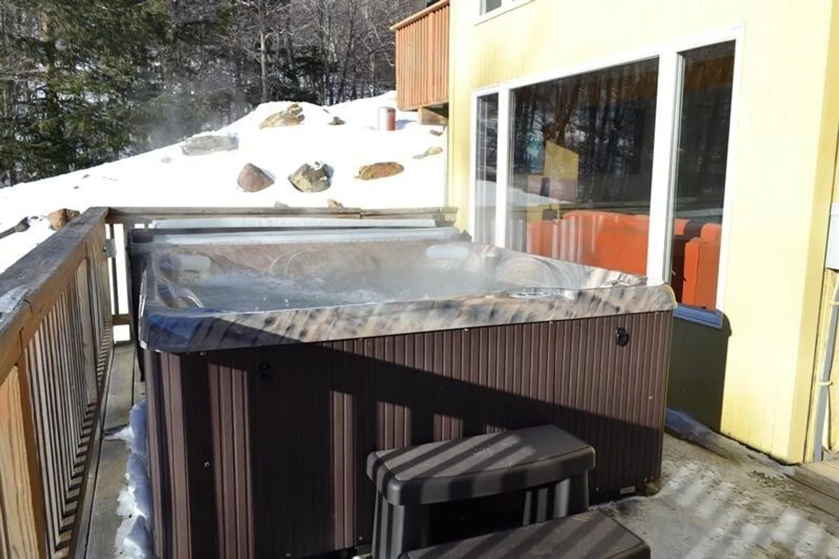 This image captures an inviting hot tub set amidst a snowy landscape, framed by surrounding trees and large windows leading back inside. The tranquil winter scenery emphasizes relaxation, with the hot tub clearly the centerpiece for unwinding after a day outdoors.