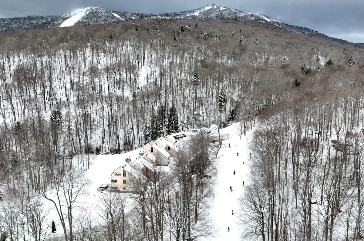 A wide exterior vista of a snow-blanketed landscape, distant mountains glowing at sunrise, a wooded area, and scattered cabins. The mood is peaceful, suggesting the serenity found in nature after a day on the slopes.