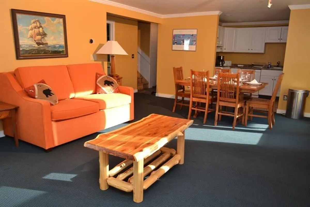 The image displays a cozy, warmly lit living and dining area featuring an inviting orange sofa, a wooden coffee table, and a dining table with chairs. A painting of a ship adds a personal, artistic touch to the space. The setting conveys a sense of comfort and homeliness, ideal for relaxing or socializing after a day of outdoor adventure.