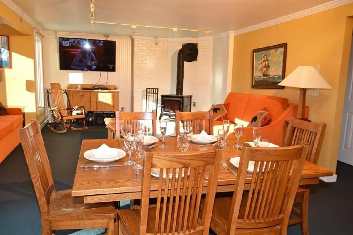 A warmly lit interior reveals a wood-burning stove, comfortable orange sofa, dining table, and television. The layout encourages conversation and togetherness, making it ideal for family dinners or cozy movie nights after a day outdoors.